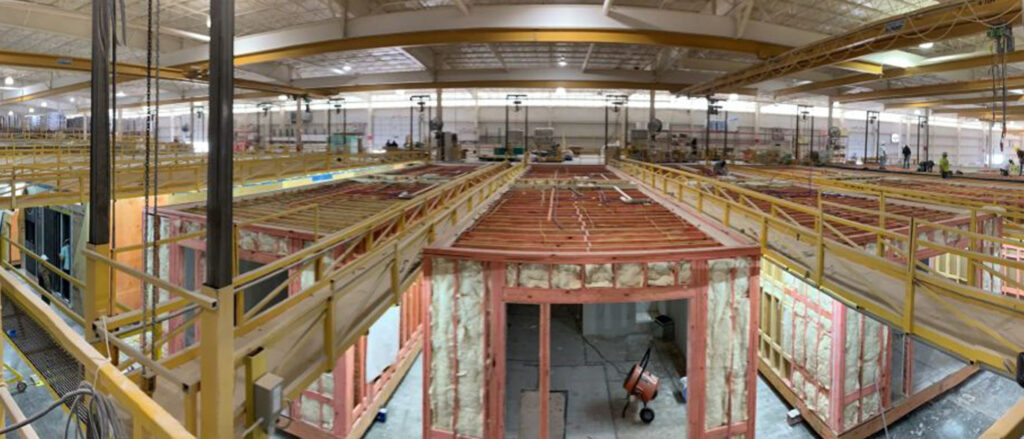 Manufactured homes in various stages of construction inside a large manufactured home facility, with exposed framing, insulation, and overhead walkways visible.
